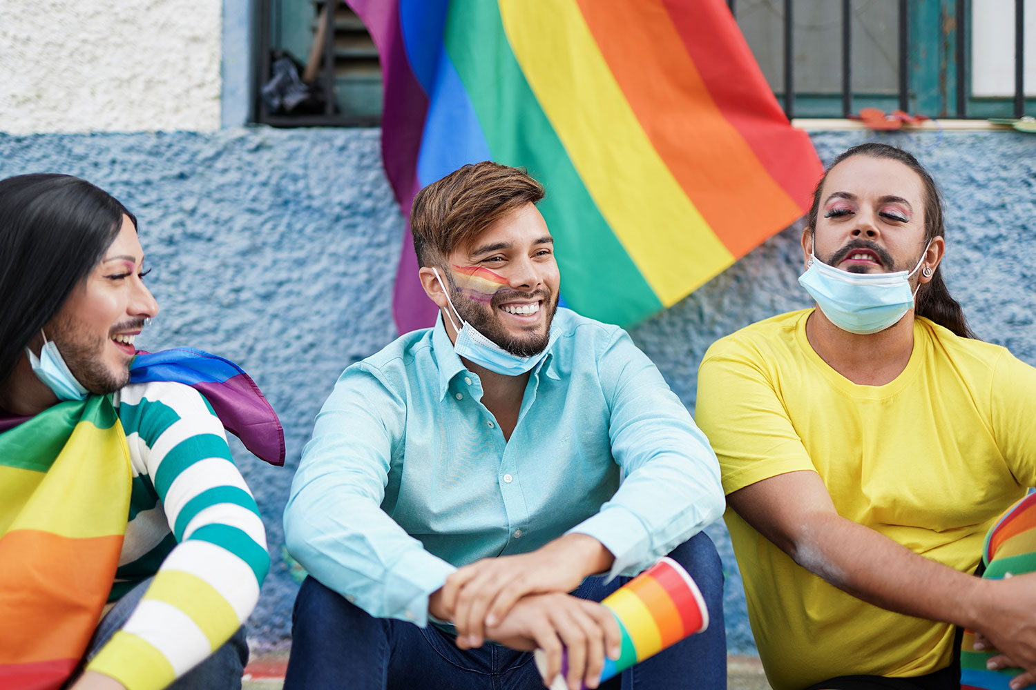 three young adults smiling wearing the pride flag on socks shirts face paint with large pride flag behind them